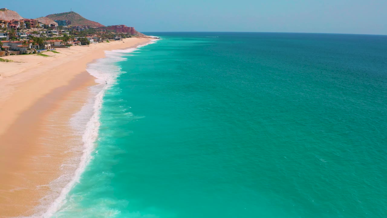 Aerial frame at Dreams Beach showing the coast receding into the distance, foamy waves along golden sand, blue-green water, and no visible people.