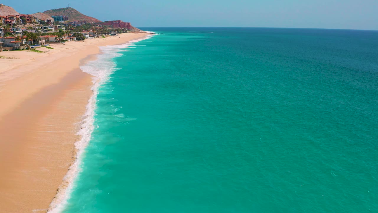 Aerial beach edge with white surf entering from the lower left and open ocean to the right.