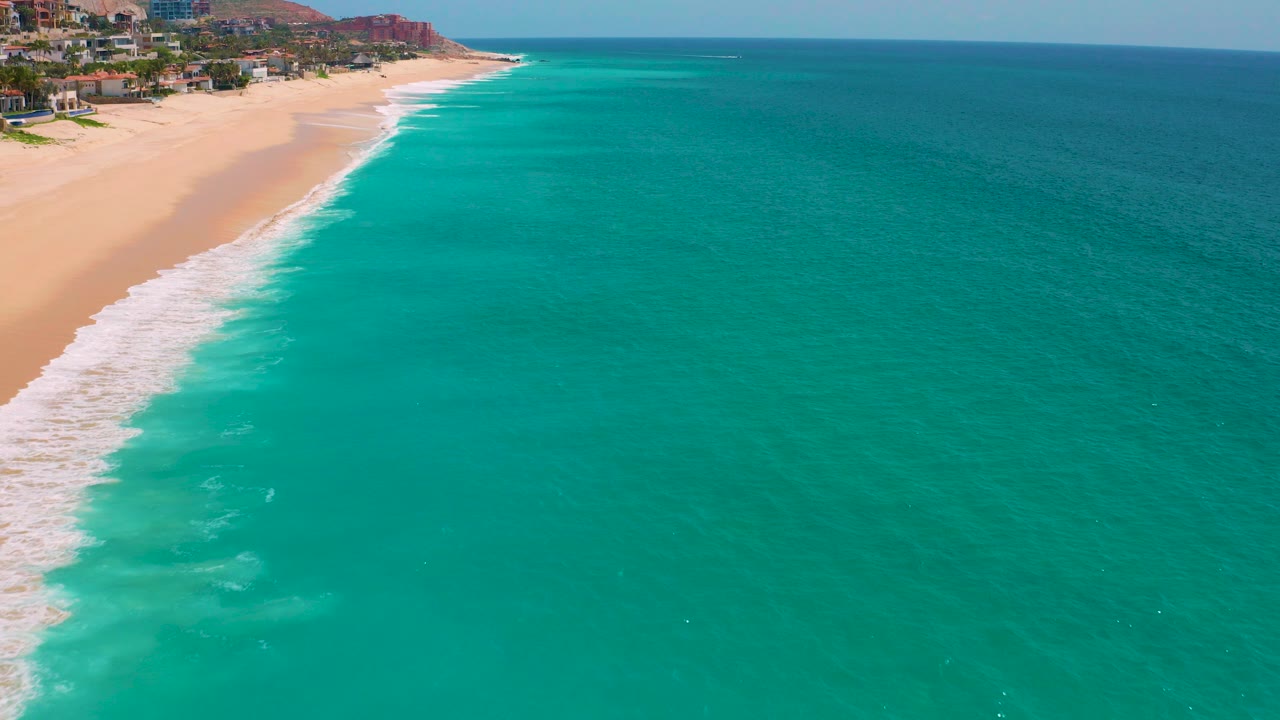 Aerial frame over Dreams Beach in Baja California Sur, Mexico, with turquoise water, a white surf band along the left beach edge, coastal buildings, distant reddish hills, and no visible people.
