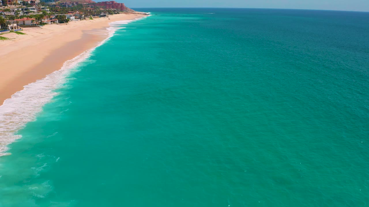 Aerial frame over Dreams Beach in Baja California Sur, Mexico, with most of the image filled by blue-green water, a narrow sandy shore and breaking surf at left, hillside development near the top, and no visible people.