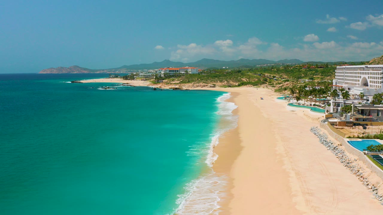 Opening frame over Dreams Beach in Baja California Sur, Mexico, with turquoise water at left, a pale sand shoreline through the center, resort buildings at right, distant hills, and no visible people.