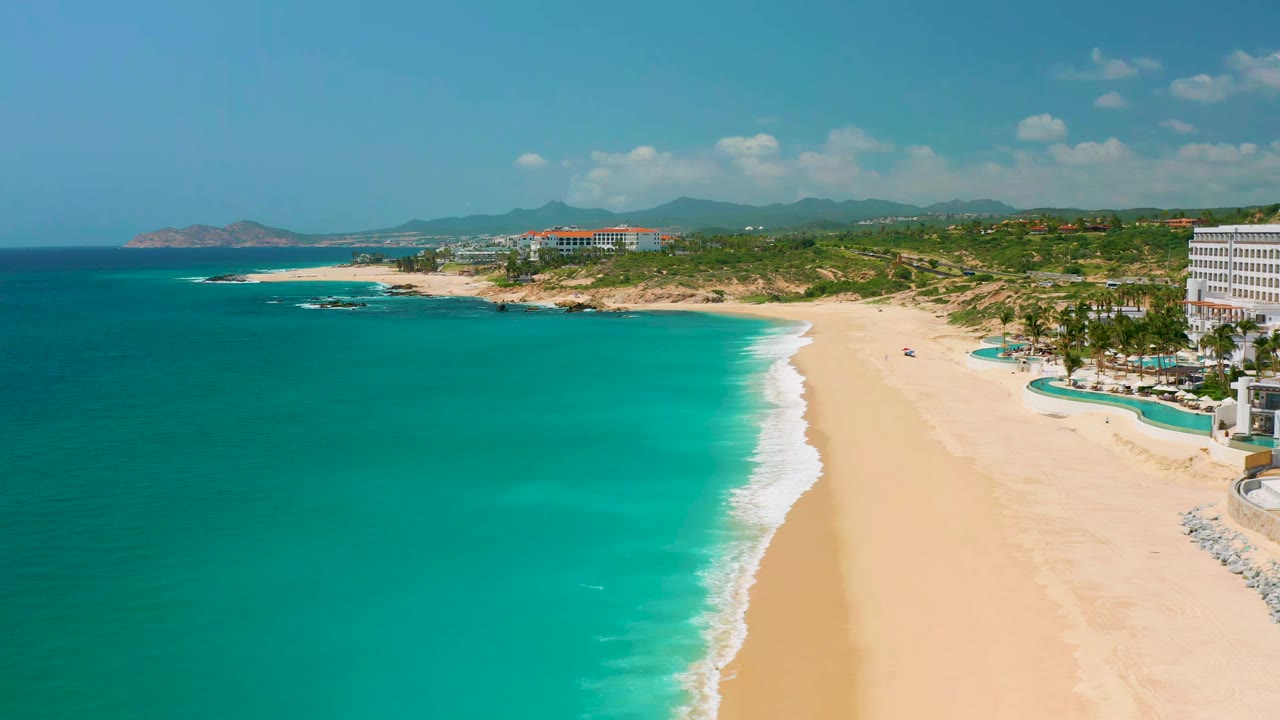 Wide aerial frame of Dreams Beach showing blue-green water, a curving white surf line, pale sand, low hills beyond the coast, resort-area buildings, and no visible people.