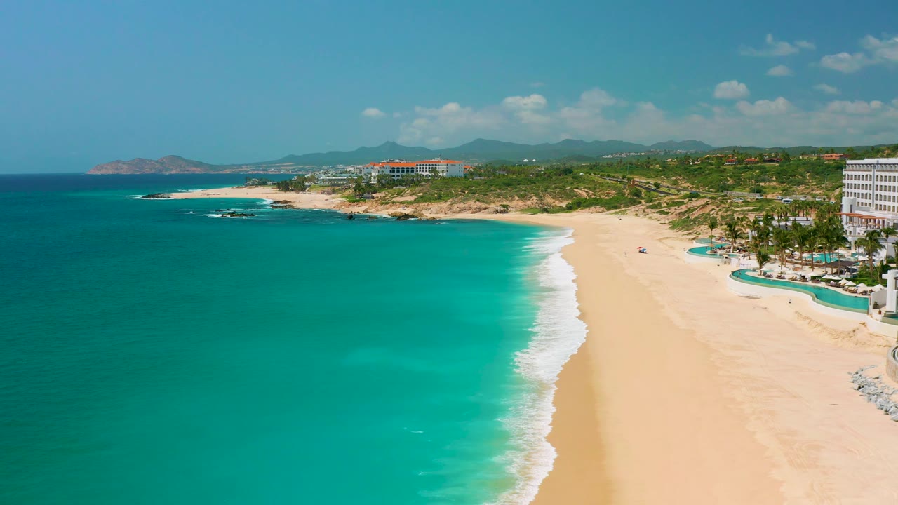 Aerial view of Dreams Beach with more open turquoise water in front, a bright sand strip to the right, white surf, palms near the resort edge, distant hills, and no visible people.