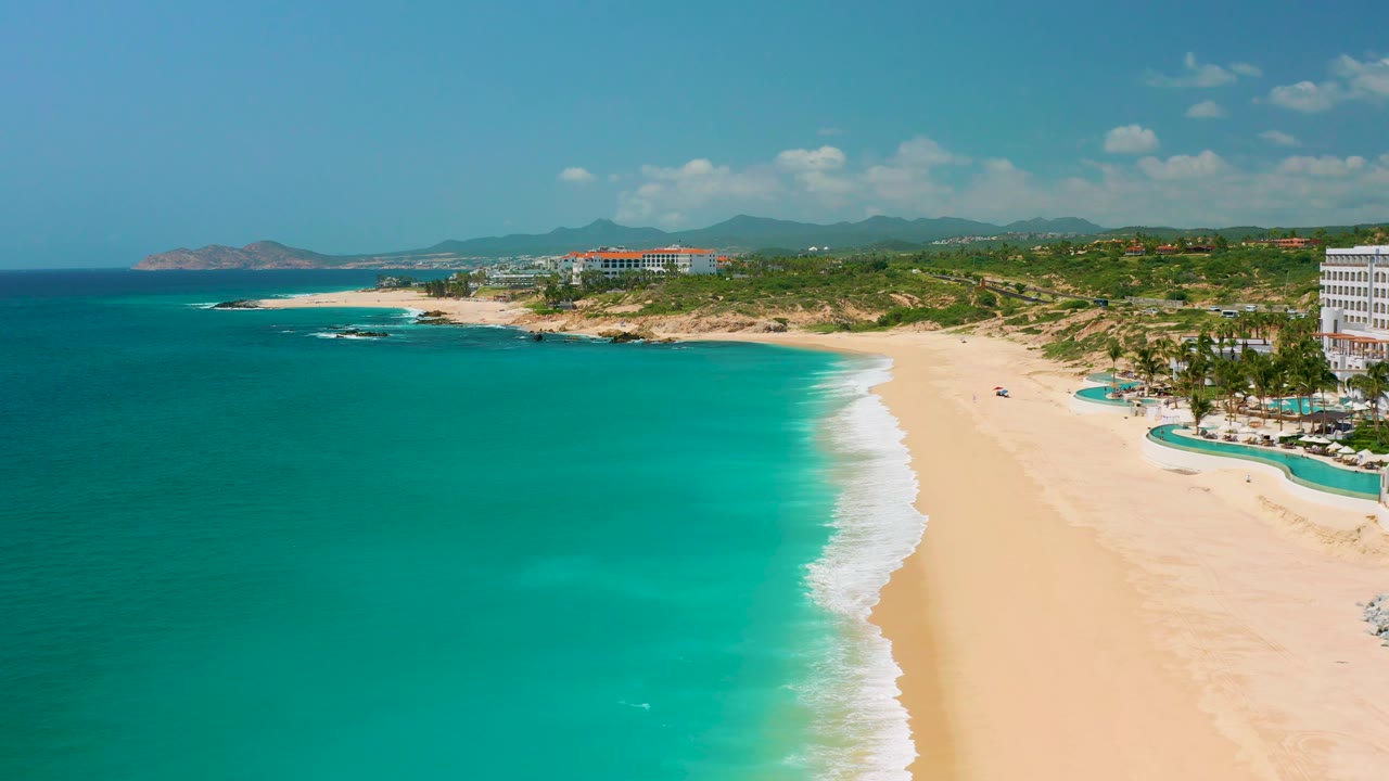 Aerial frame over Dreams Beach with the ocean taking most of the left side, a narrow white break of waves, sandy beach to the right, resort-area buildings inland, and no visible people.