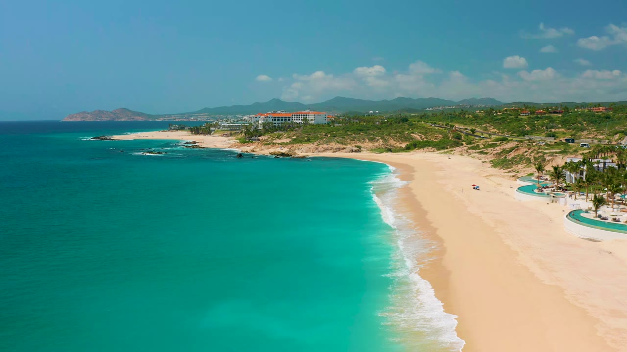 Aerial view of Dreams Beach in Baja California Sur, Mexico, showing a curved white surf line between clear blue-green water and open sand, with no visible people.