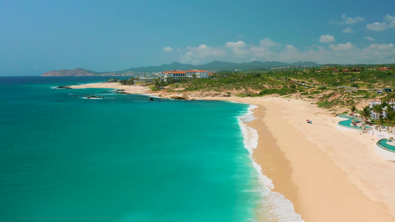 Aerial frame over Dreams Beach in Baja California Sur, Mexico, with turquoise water, pale sand, a rocky point in the distance, resort-style buildings, and no visible people.