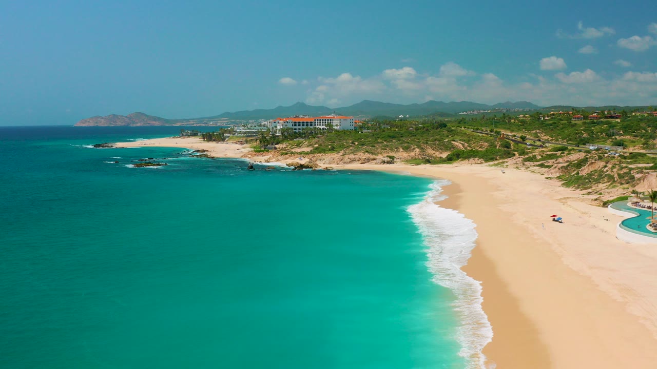 Opening aerial view at Dreams Beach in Baja California Sur, Mexico, with turquoise water on the left, pale sand on the right, white surf along the shore, distant buildings and hills, and no visible people.