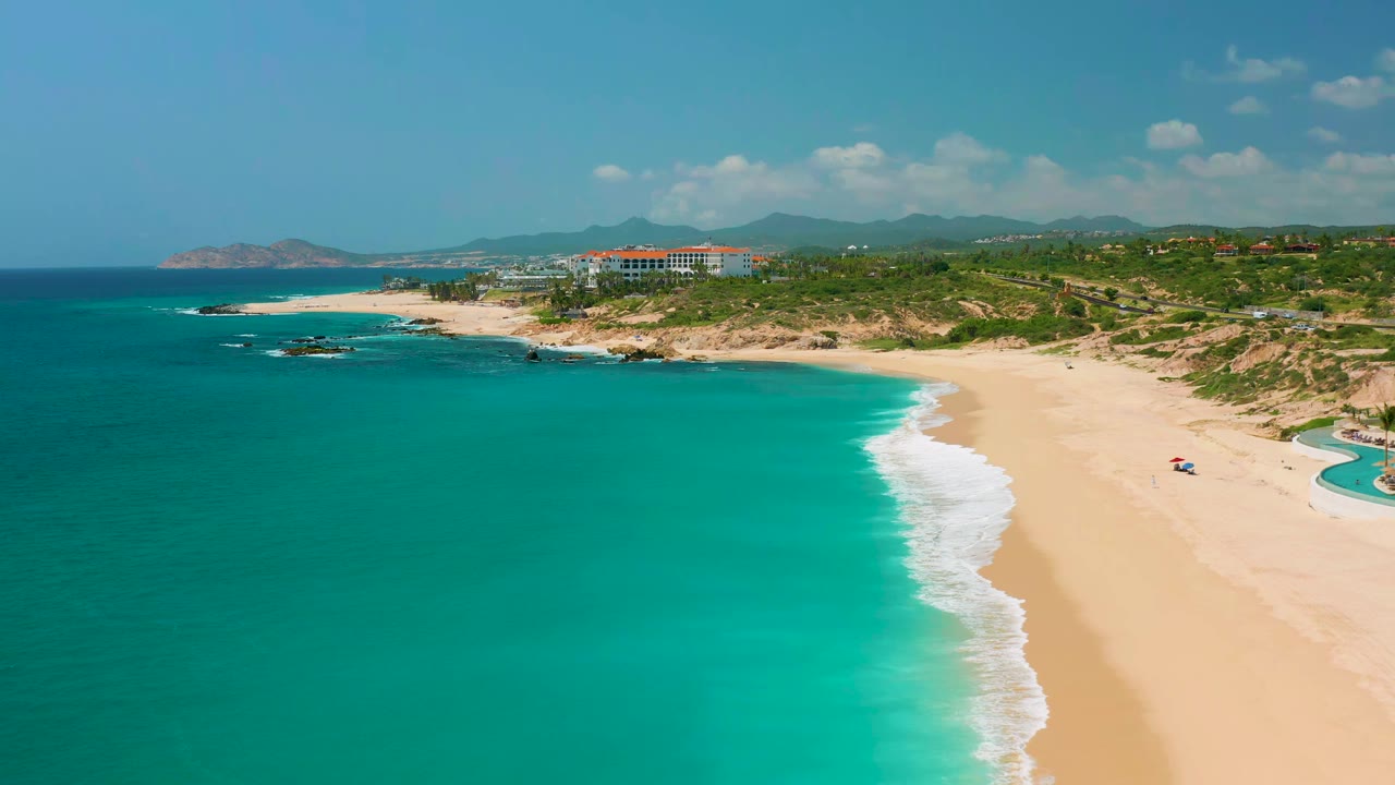 One second into the clip, the aerial view holds over Dreams Beach with a longer sweep of turquoise water, a curving white wave line, open sand, green coastal terrain, and no visible people.