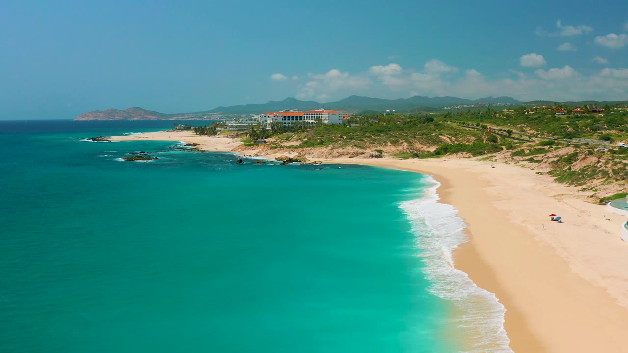 Wide aerial of ocean and beach with rocky points, surf, dunes, buildings, mountains, and clouds.