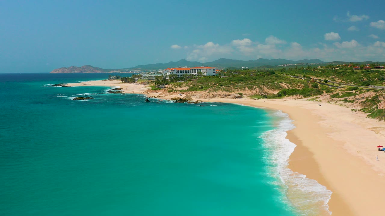 Aerial frame over Dreams Beach in Baja California Sur with turquoise water in the foreground, a pale sand shoreline on the right, rocks to the left, and resort buildings beneath distant mountains.