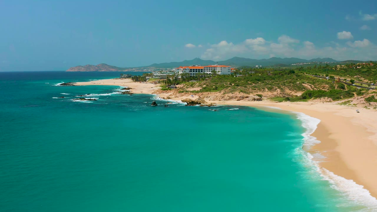 Wide aerial view of Dreams Beach in Baja California Sur, Mexico, showing a broad turquoise bay, pale sand along the right edge, low rocks near the surf, resort-area buildings, and no visible people.