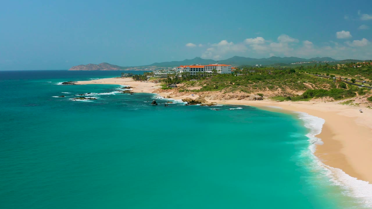 Aerial view along Dreams Beach in Baja California Sur, Mexico, with the turquoise sea filling most of the frame, a bright sand curve on the right, white surf at shore, and no visible people.