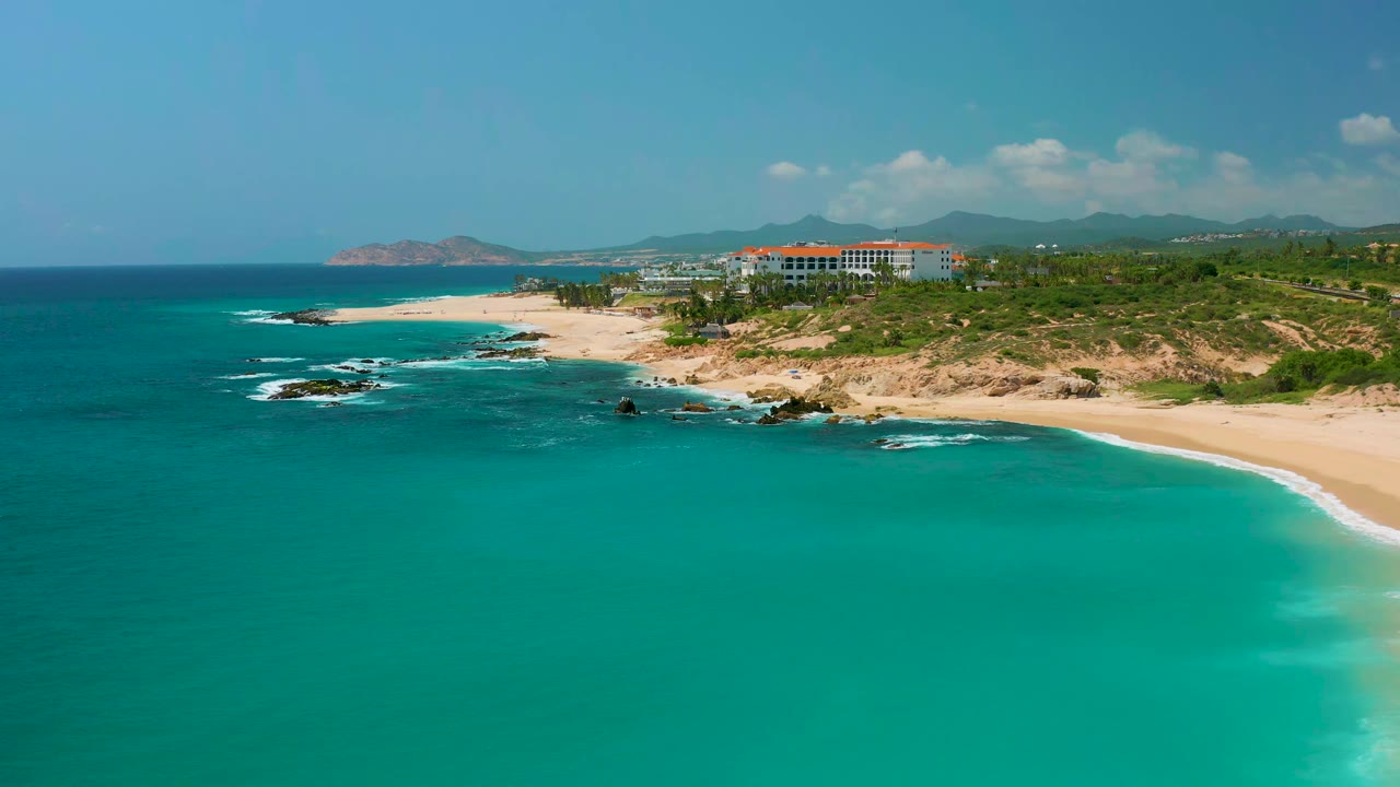 Wide aerial coastline with blue-green sea, bright beach, dark rocks, and resort buildings inland.