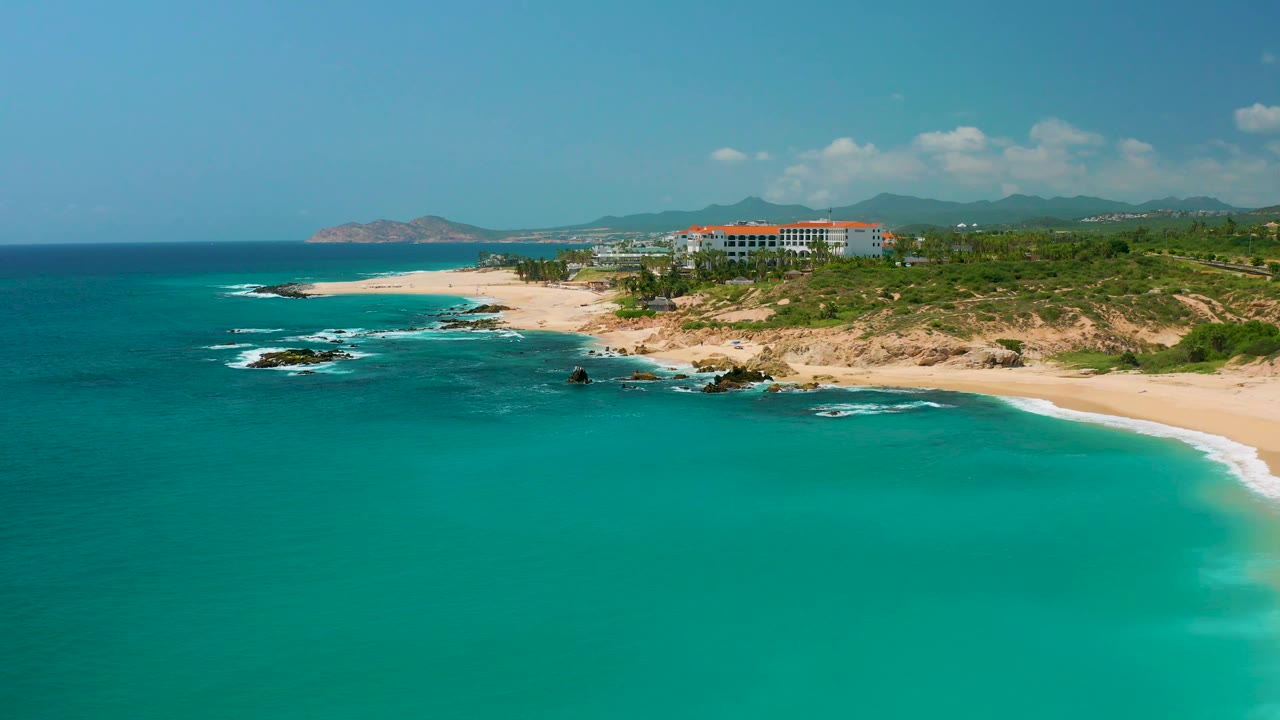 Dreams Beach aerial view with calm turquoise water dominating the lower frame, rocky points interrupting the sand, a red-roofed resort near the upper right, and no visible people.