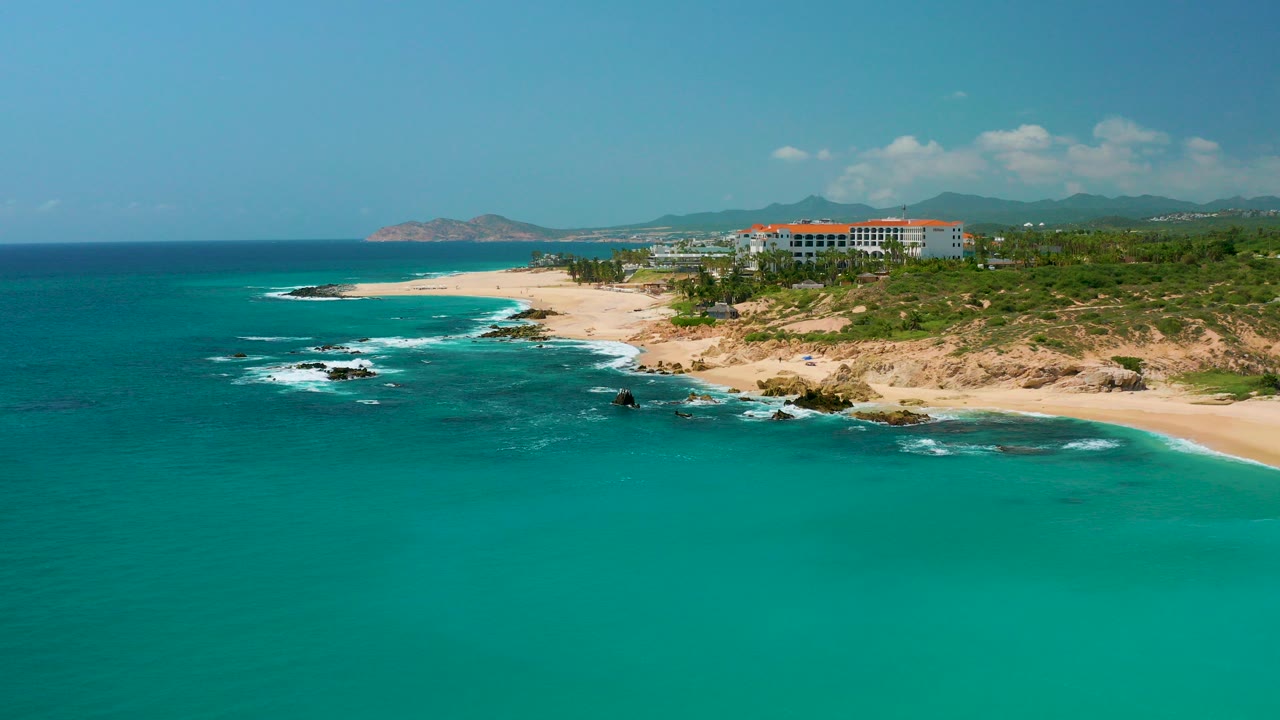 Opening frame over Dreams Beach in Baja California Sur, Mexico, with turquoise water in the foreground, white surf on dark rocks, sandy shoreline, resort buildings, distant mountains, and no visible people.
