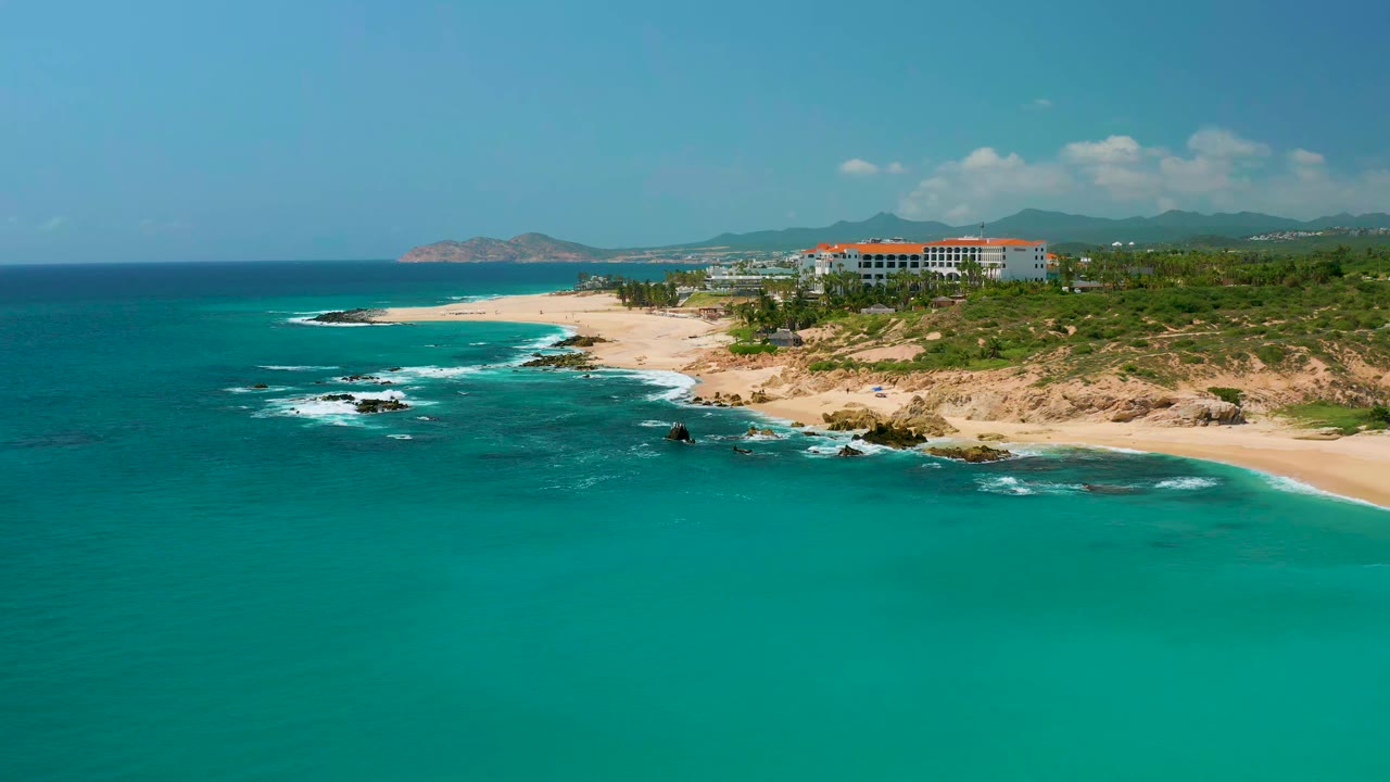 Wide coastal aerial with surf breaking near rocks and resort buildings above the beach.