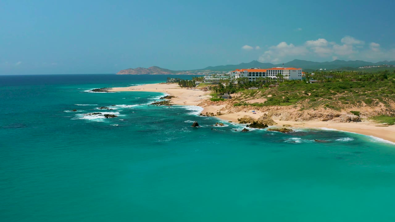 At 0:02, the aerial view keeps Dreams Beach framed with open turquoise water below, a curved sandy shore, dark rocks with white water, resort buildings inland, mountains behind them, and no visible people.
