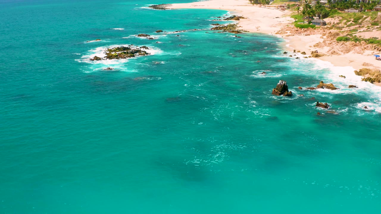 Turquoise sea with white surf around rocks and sandy shoreline beyond.