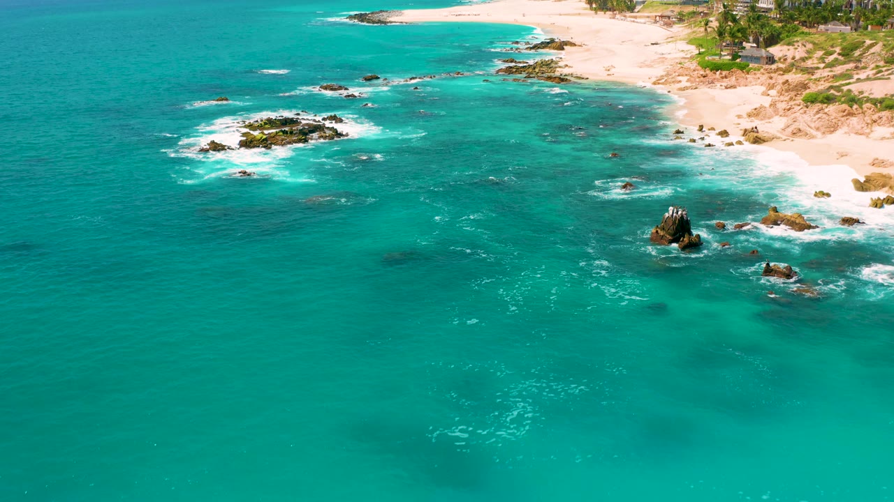 Aerial frame over Dreams Beach showing more open water in front, rocks and surf near the right side, bright sand and palms along the top edge, and no visible people.