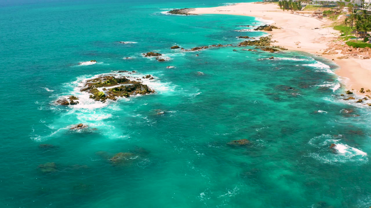 Aerial view of turquoise ocean, rocks, surf, and sandy beach.