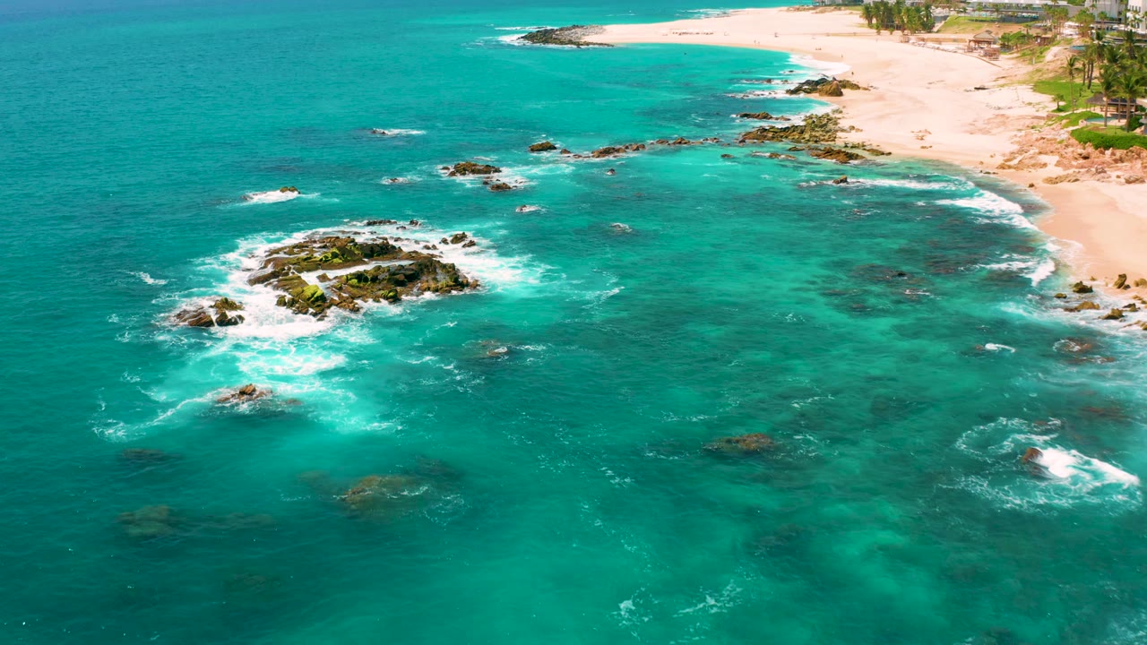 Aerial view at Dreams Beach with white waves wrapping the central rock formation, shallow turquoise water, beach at the top of frame, and no visible people.