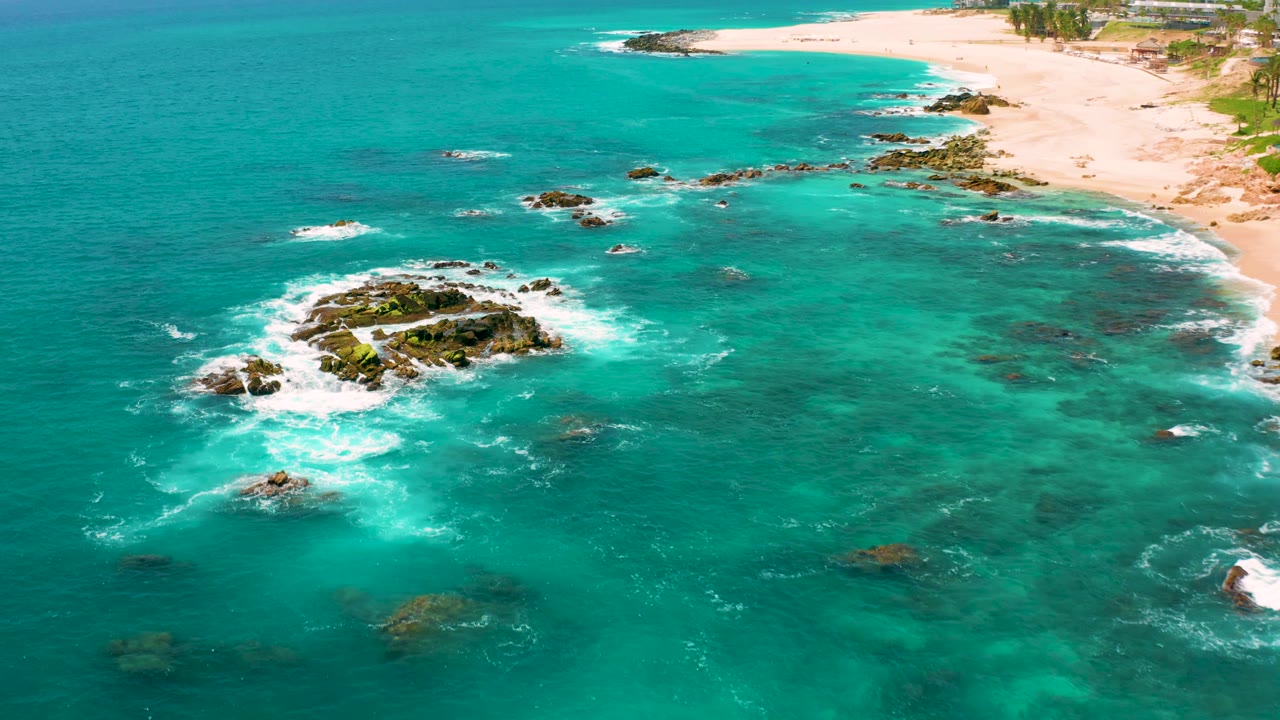 Drone frame over Dreams Beach showing a wider spread of turquoise ocean, scattered rocks, white surf, pale sand, and no visible people.