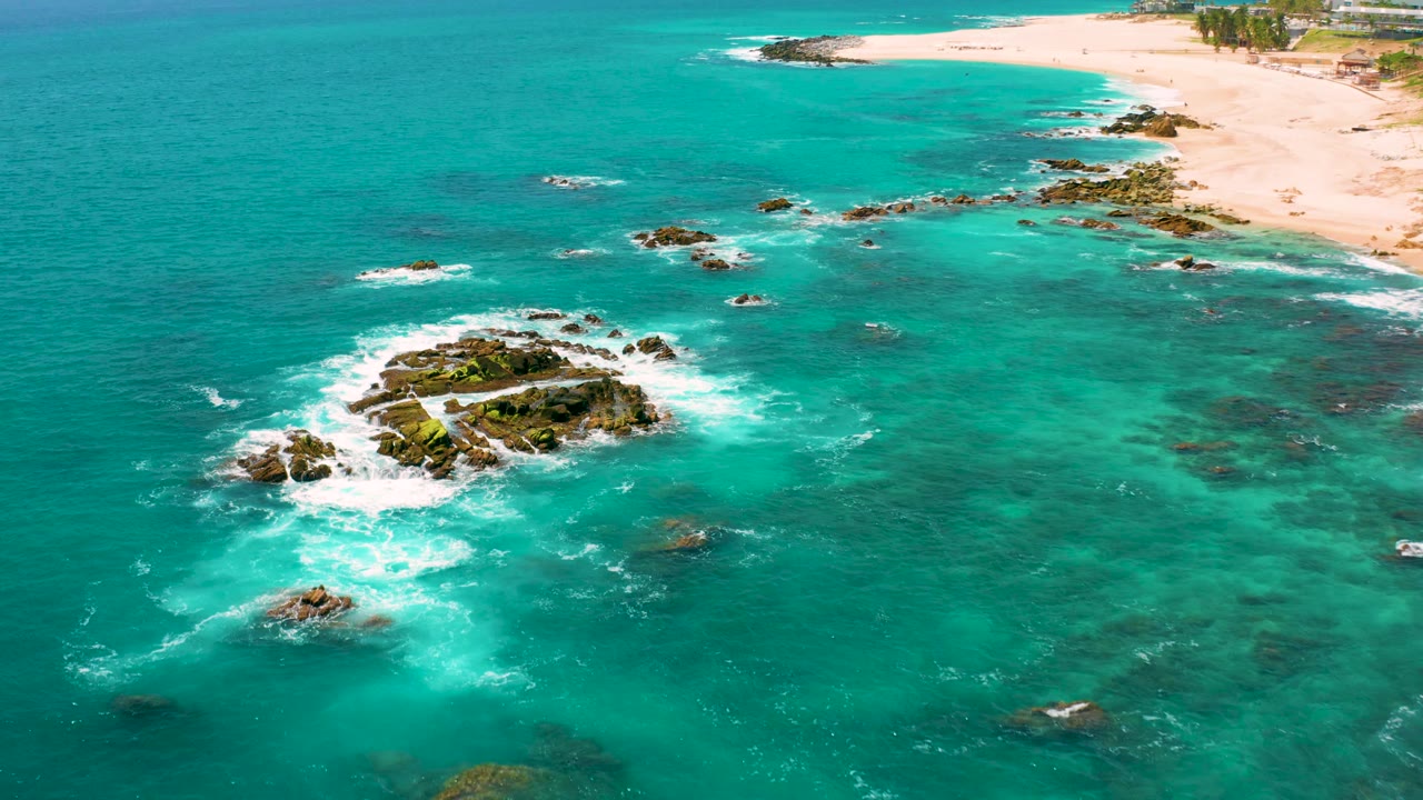 Drone view at Dreams Beach with the main rock outcrop lower left of center, foamy water around it, turquoise sea across the frame, and no visible people.