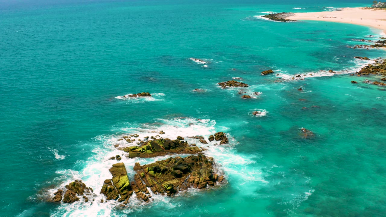 Aerial frame at Dreams Beach, Baja California Sur, showing turquoise water, white surf around foreground rocks, a distant sandy beach, and no visible people.