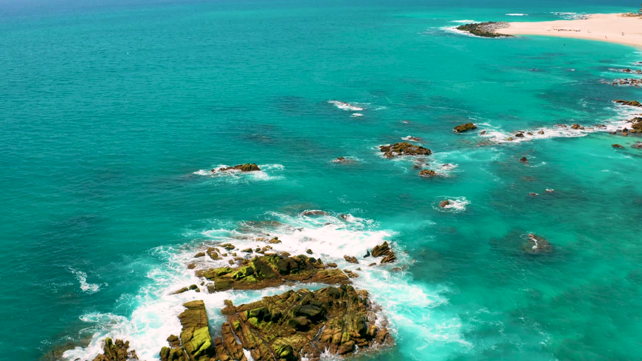 Aerial frame at Dreams Beach, Baja California Sur, showing more open turquoise water, scattered rocks, surf bursting near the lower rock edge, and no visible people.