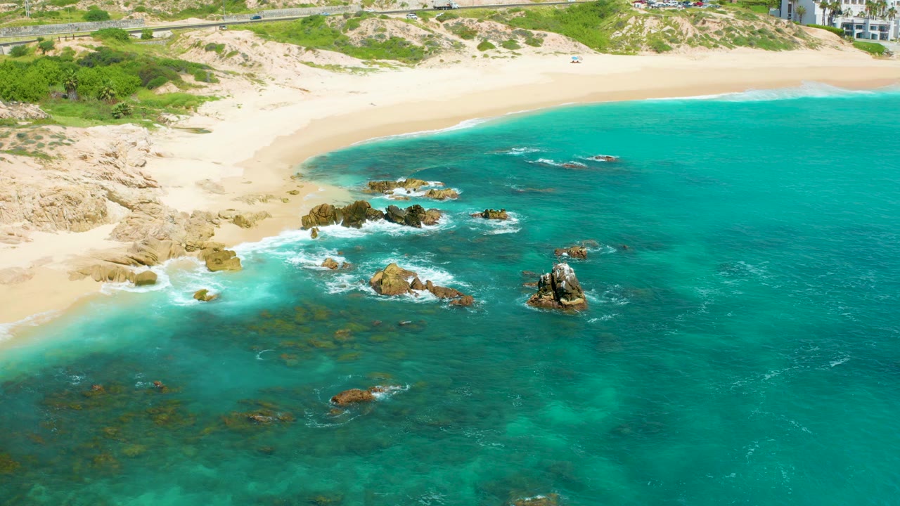 Aerial frame over Dreams Beach in Baja California Sur, Mexico, showing turquoise water washing around scattered shoreline rocks beside pale sand, with no visible people.