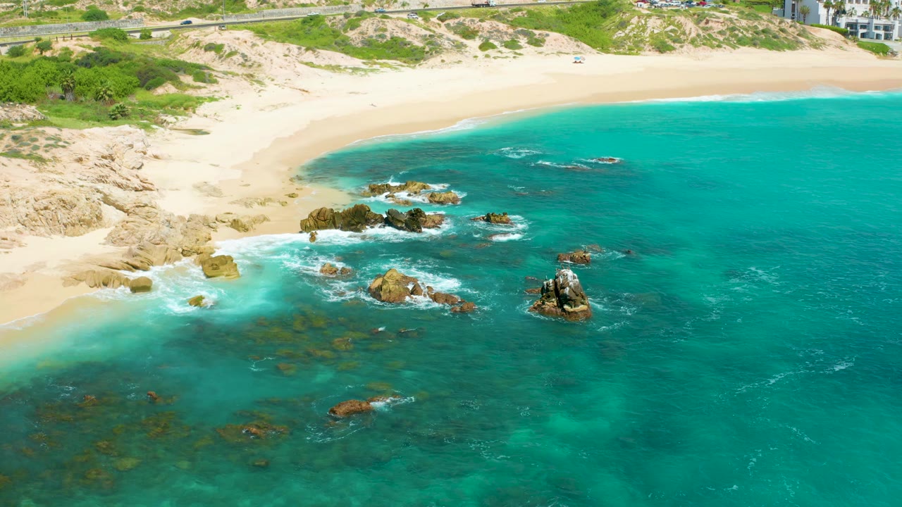 Aerial frame over Dreams Beach in Baja California Sur, Mexico, showing a curving sand beach above rocky turquoise shallows, with no visible people.