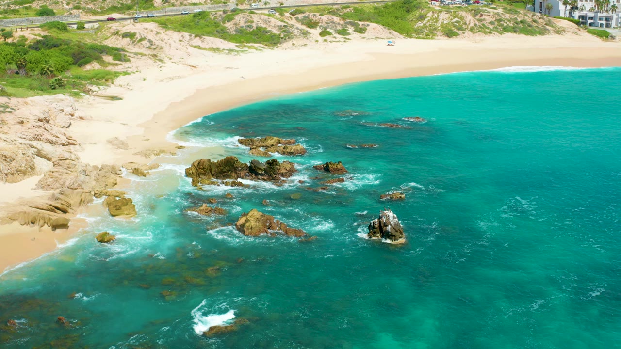 Aerial frame over Dreams Beach in Baja California Sur, Mexico, showing turquoise water, scattered shoreline rocks, pale sand, dunes, a road, distant hotel buildings, and no visible people.