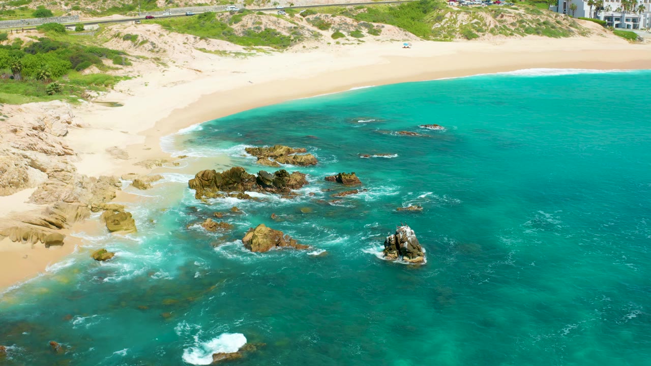 High aerial frame over Dreams Beach showing more open turquoise water to the right, tan rocks near the surf line, a smooth sandy beach, coastal vegetation, a road, hotel buildings, and no visible people.