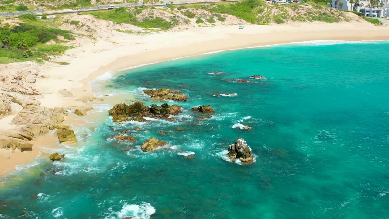 Aerial frame from Dreams Beach with white surf spreading across darker shallow water, large tan rocks in the lower center, the beach sweeping across the top, and no visible people.