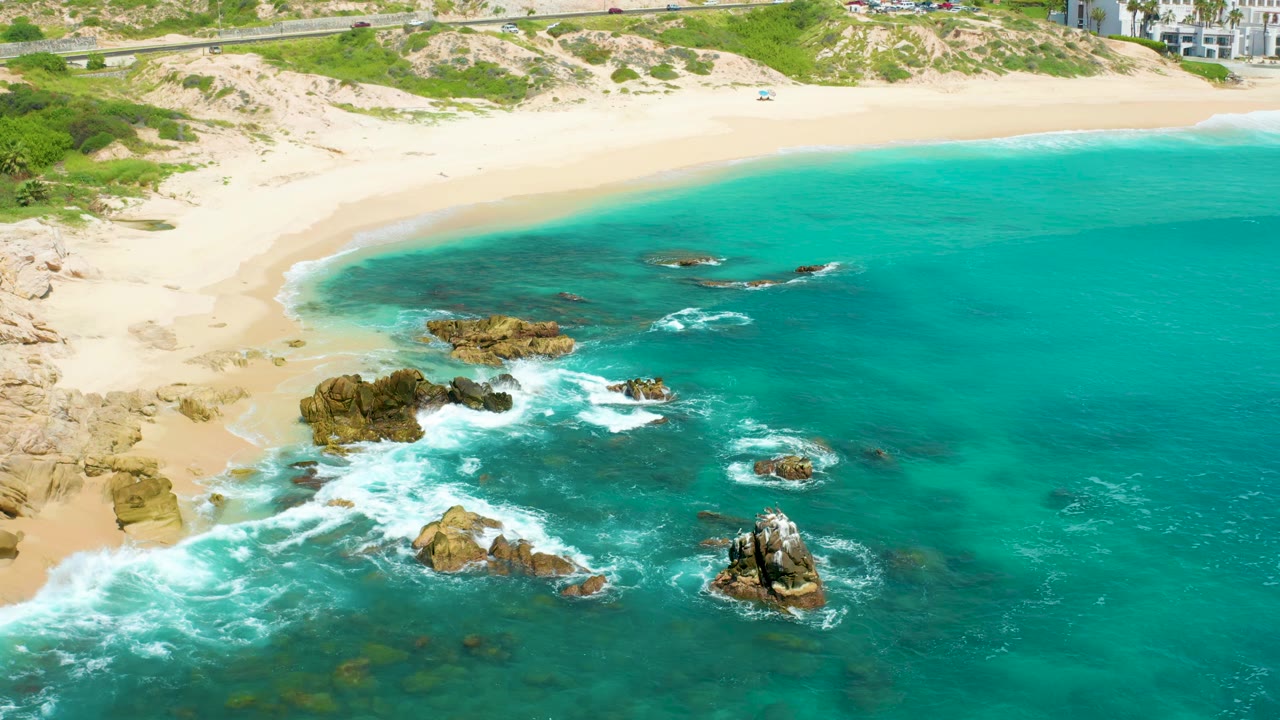 Blue-green surf curls around offshore rocks near a wide sandy shoreline.