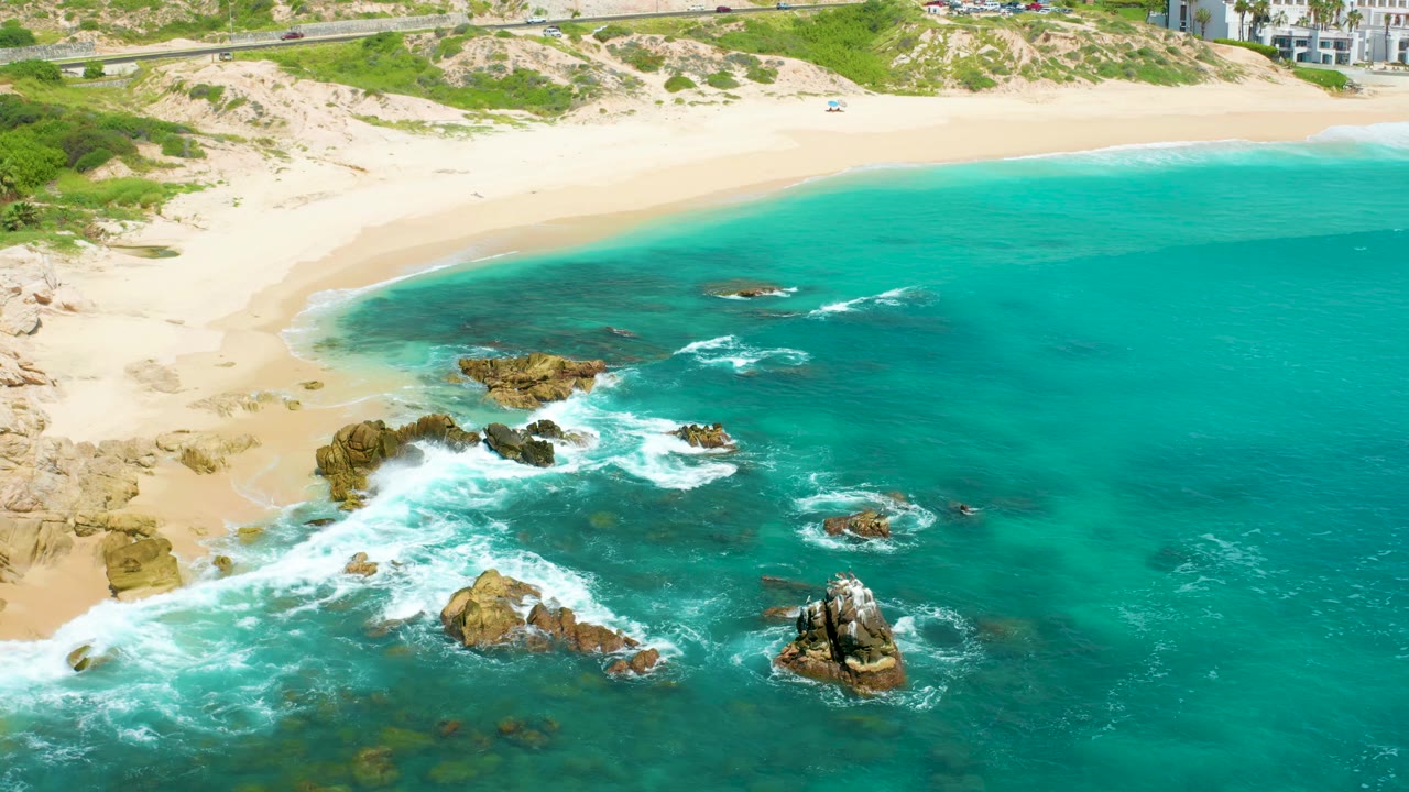 Aerial frame from Dreams Beach showing whitewater spreading over dark rocks in turquoise shallows, tan sand and low dunes along the top edge, and no visible people.