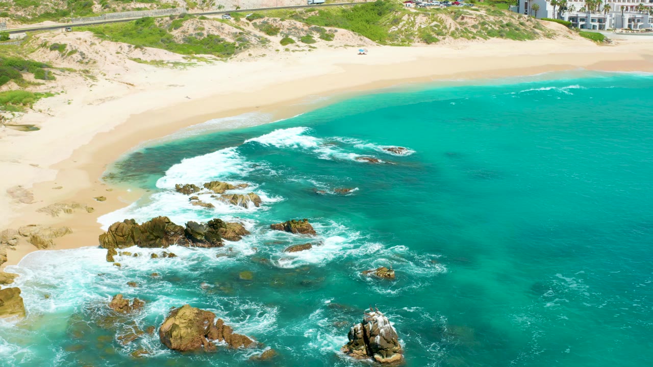 Aerial coastline with waves breaking over rocks beside a sandy beach.
