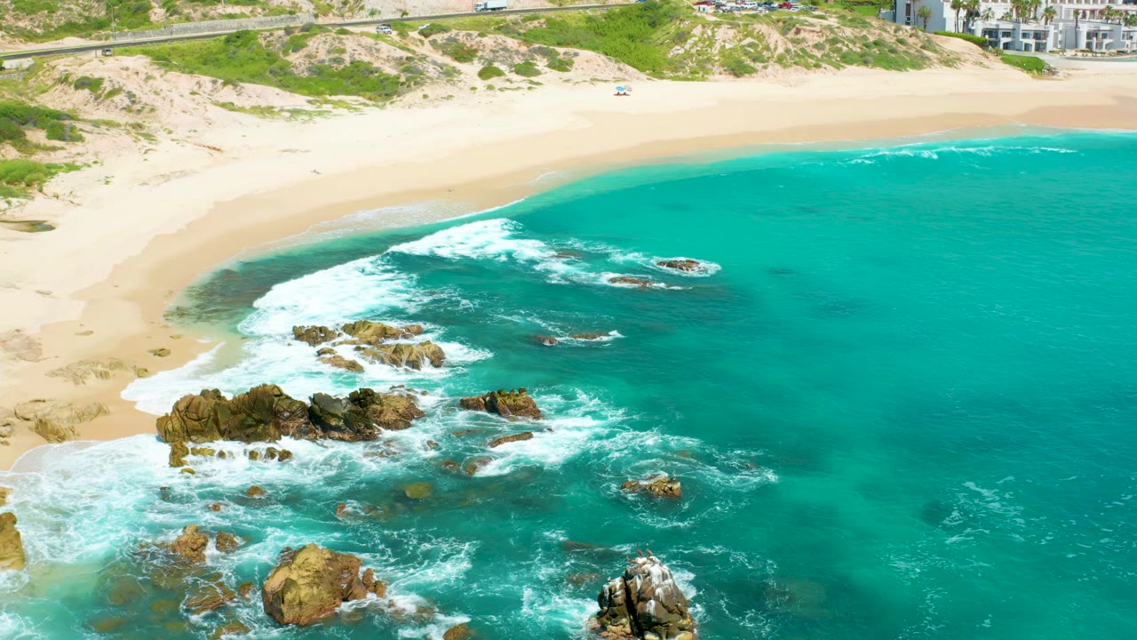 High aerial frame of Dreams Beach in Baja California Sur with a curved sandy shoreline, white surf crossing near rocks, open turquoise water, and no visible people.