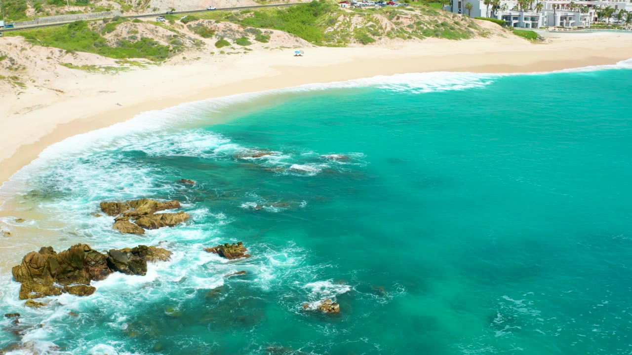 At 0:01, the aerial frame shows white foam spreading across the rock cluster below the sand and dunes at Dreams Beach, with no visible people.