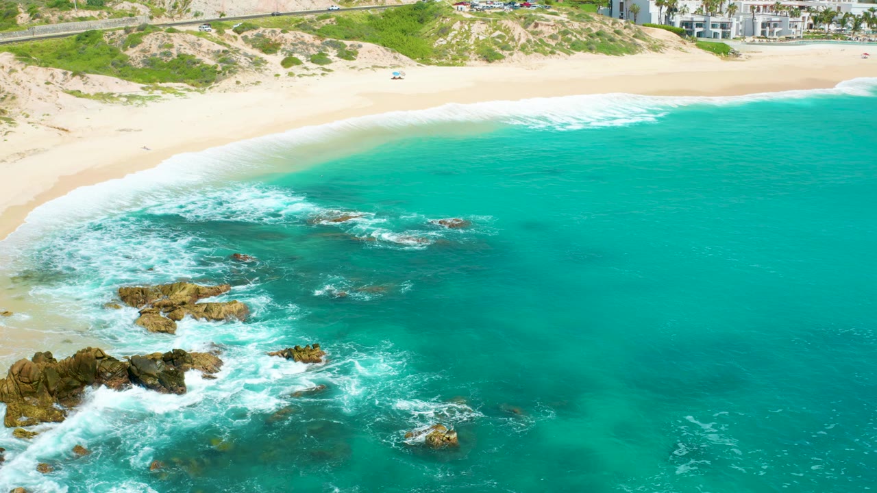 Aerial frame at Dreams Beach with more open turquoise water in view, surf curling around scattered rocks, and the beach and buildings along the top edge, with no visible people.
