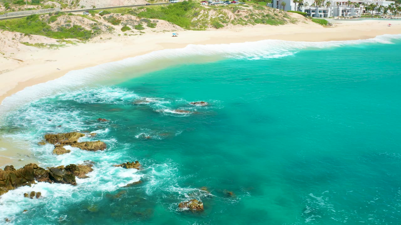 Mid-clip aerial view at Dreams Beach showing a rock formation in the lower left, white water washing around it, and a wide band of teal ocean, with no visible people.