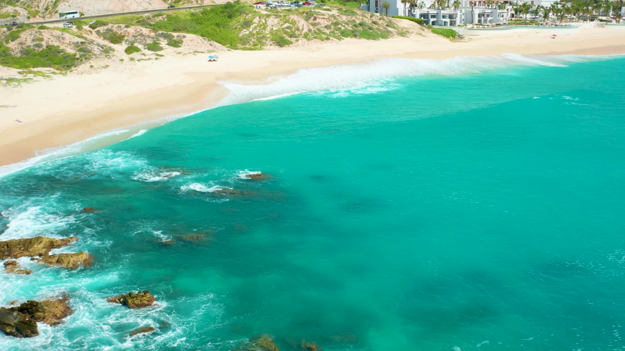 Aerial frame over Dreams Beach, Baja California Sur, with turquoise water filling most of the view, rocks and white surf at lower left, pale sand and green dunes along the top, distant buildings, and no visible people.