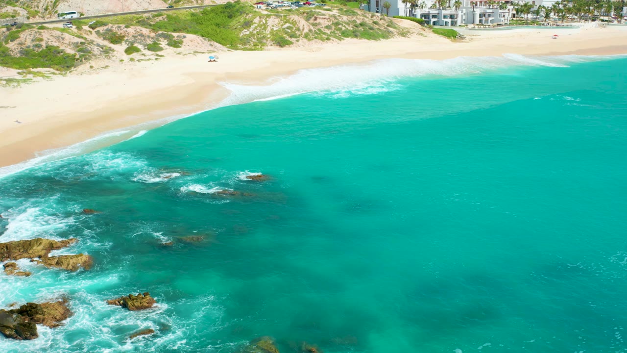 Aerial beach scene with clear water, surf over rocks, sand, dunes, and buildings.