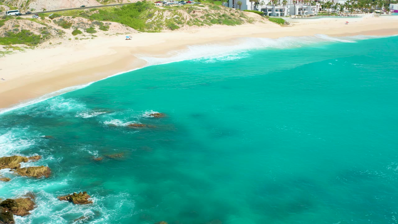Aerial frame of Dreams Beach with a long white surf line along pale sand, turquoise water and darker reef-like patches offshore, rocks at lower left, small vehicles above the dunes, buildings at the top, and no visible people.