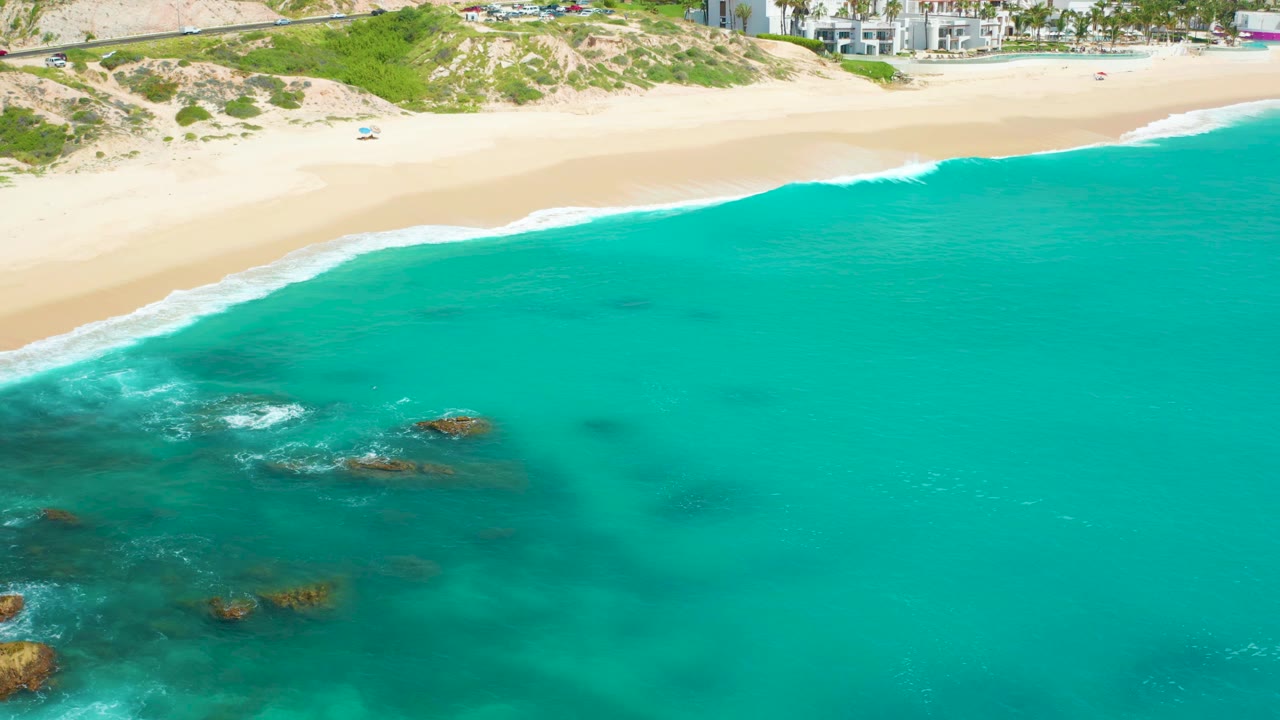 Aerial frame over Dreams Beach with submerged rock patches in the lower water, a white surf line along the sand, dune vegetation, distant buildings, and no visible people.
