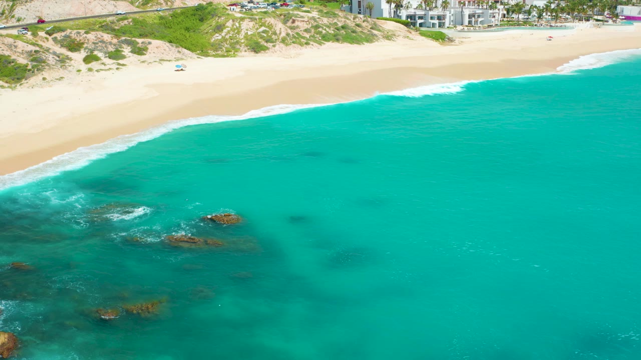 Aerial frame over Dreams Beach showing more open turquoise water to the right, rocks and foam to the left, pale sand at the top, and no visible people.