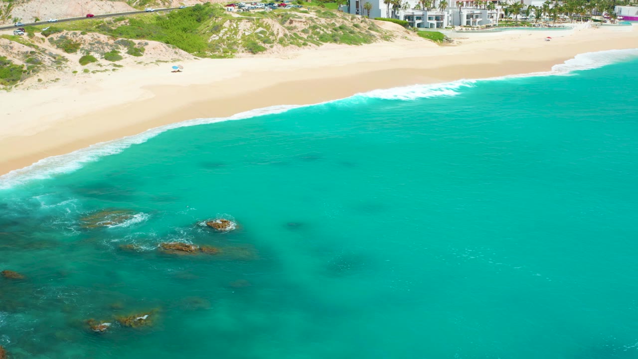 Aerial frame over Dreams Beach with small waves breaking near exposed rocks, clear turquoise water filling most of the frame, empty sand above, and no visible people.