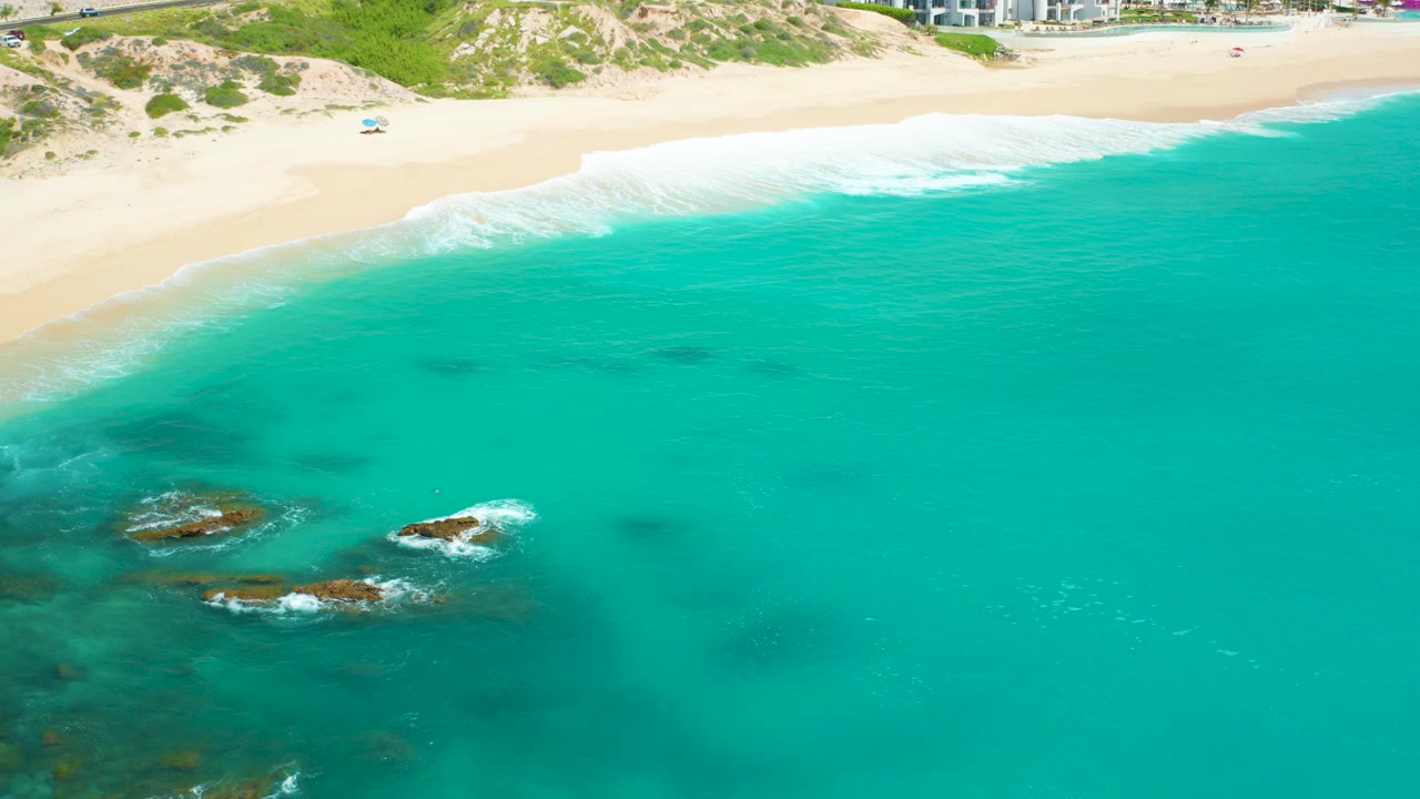 Aerial frame over Dreams Beach in Baja California Sur, with turquoise water around dark reef rocks, pale sand and low dunes at the top, and a tiny distant beachgoer near the shore.