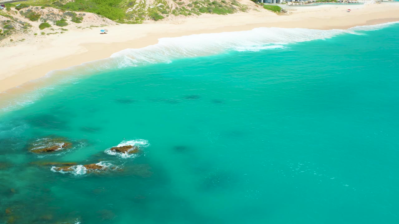 Drone-like view over Dreams Beach with open turquoise water dominating the frame, reef rocks toward the lower left, surf lines along the sand, and a tiny person visible high on the beach.