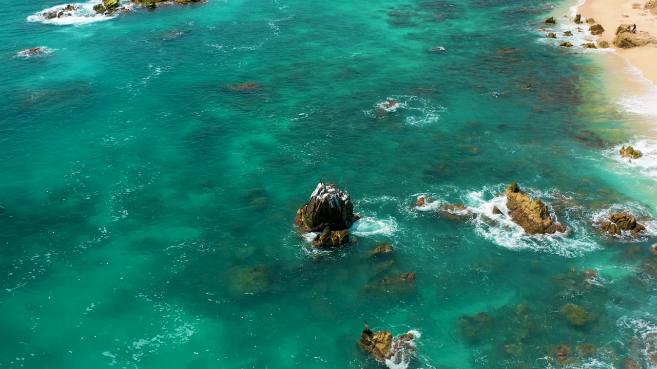 Aerial frame over Dreams Beach in Baja California Sur, Mexico, with turquoise water, scattered rock outcrops, white surf, sandy beach at upper right, and no visible people.